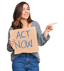 Young brunette woman holding act now banner smiling happy pointing with hand and finger to the side
