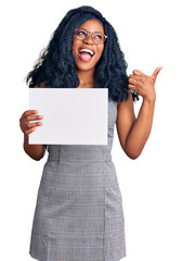 Beautiful african american woman holding blank empty banner pointing thumb up to the side smiling happy with open mouth