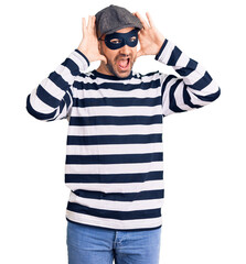 Young handsome man wearing burglar mask smiling cheerful playing peek a boo with hands showing...