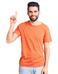Young handsome man with beard wearing casual t-shirt showing and pointing up with finger number one while smiling confident and happy.