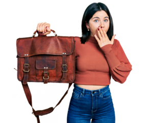 Young brunette woman with blue eyes holding leather bag covering mouth with hand, shocked and afraid for mistake. surprised expression