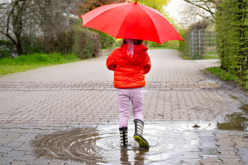 mischievous 5-year-old girl in rubber boots with red umbrella stands in rain puddle, capturing pure...