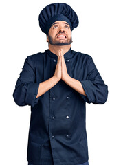 Young hispanic man wearing cooker uniform begging and praying with hands together with hope...