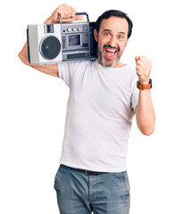 Middle age handsome man listening to music using vintage boombox screaming proud, celebrating victory and success very excited with raised arms