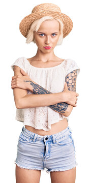 Young blonde woman with tattoo wearing summer hat skeptic and nervous, disapproving expression on face with crossed arms. negative person.