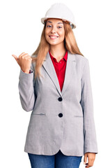 Beautiful young woman wearing architect hardhat smiling with happy face looking and pointing to the side with thumb up.