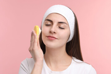 Young woman with headband washing her face using sponge on pink background
