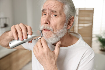 Senior man trimming mustache with electric trimmer in bathroom - obrazy, fototapety, plakaty