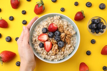 Woman holding bowl of tasty oatmeal with strawberries, blueberries and walnuts on yellow background, top view