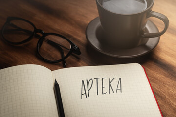 A handwritten inscription "Apteka" on a grille of an open notebook on a wooden countertop, next to a pencil, a cup with coffee and glasses, a flash of light. (selective focus), translation: Pharmacy