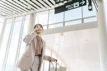 スマホで電話しながら空港・駅・ビル・公共交通施設でスーツケースを持って歩く女性（海外出張・徒歩）
