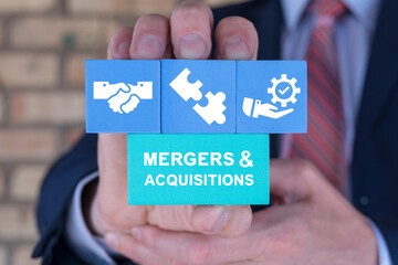 Business man holding blocks with icons sees text: MERGERS AND ACQUISITIONS. Concept of business mergers and acquisitions development model. Share acquisition, asset business acquisition, amalgamation.