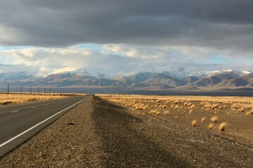 Panoramic shot of a smooth asphalt road running along a power line in the picturesque autumn steppe towards the snow-capped mountains.