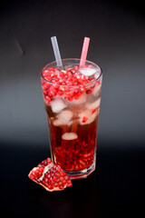 Refreshing pomegranate juice with ice and seeds in a tall glass on a black background, next to a broken fruit.