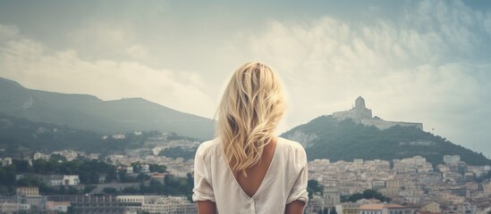 The blonde female gazes at the cityscape from a hilltop, marveling at the urban sprawl below. - obrazy, fototapety, plakaty