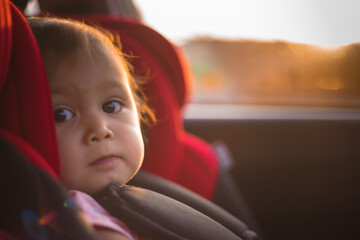 Asian Toddler Girl seats peacefully in her red carseat. She is holding her pink blanket while sucking on her thumb during sunrise or sunset. While looking at the camera.