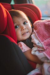 Asian Toddler Girl seats peacefully in her red carseat. She is holding her pink blanket while sucking on her thumb during sunrise or sunset. While looking at the camera.