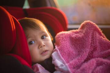Asian Toddler Girl seats peacefully in her red carseat. She is holding her pink blanket while sucking on her thumb during sunrise or sunset. While looking at the camera.