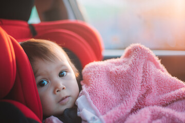 Asian Toddler Girl seats peacefully in her red carseat. She is holding her pink blanket while sucking on her thumb during sunrise or sunset. While looking at the camera.