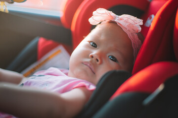 Asian Toddler Girl seats peacefully in her red carseat. On her head, she wears a pink headband with a flower on it all happening during sunrise or sunset. While looking at the camera.