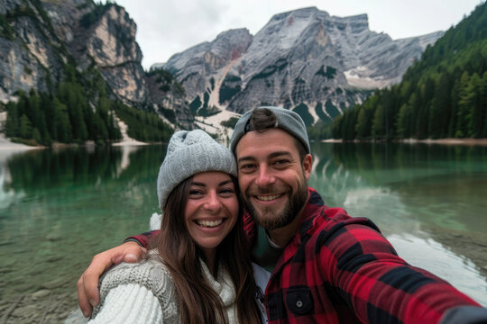 A young couple taking a selfie photo on Lake Braies in Dolomites. The man is wearing a red flannel shirt and grey beanie, the woman wears a white sweater with gray fur hat smiling at the camera