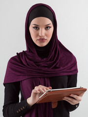 Beautiful and modern young Arabic businesswoman in hijab using a tablet computer while posing on light background and smiling at the camera. Business diversity concept, Muslim lady. - 781035715