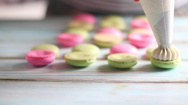 Woman pastry chef making pastel pink and green macaroons dessert. Confectioner squeezing cream filling from pastry bag to freshly baked macaron