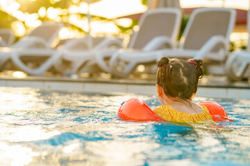 Little preschool girl with protective swimmies playing in outdoor swimming pool by sunset. Child learning to swim in outdoor pool, splashing with water, laughing and having fun. Family vacations. - obrazy, fototapety, plakaty