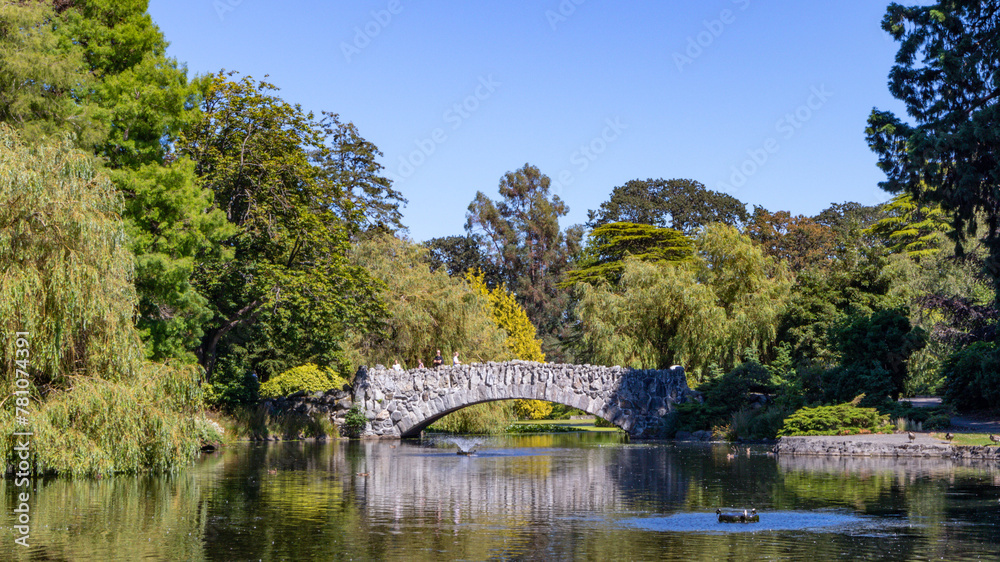 Sticker Bridge spans a tranquil pond, surrounded by trees against a clear blue sky