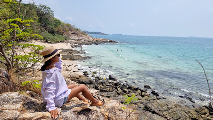 Koh Samet Island Thailand, Asian Thai women with a hat sitting on a rock looking out over the bay with a tropical beach and a blue ocean