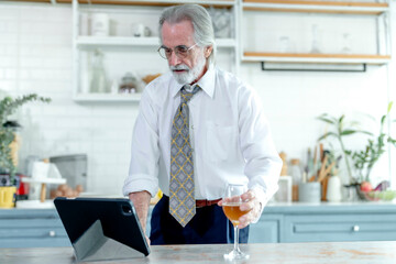Elegant senior businessman in a white shirt takes a break with a drink, using a tablet in a modern...