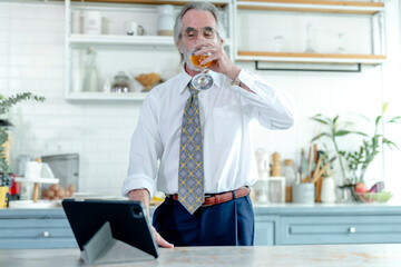 Elegant senior businessman in a white shirt takes a break with a drink, using a tablet in a modern...