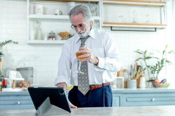 Elegant senior businessman in a white shirt takes a break with a drink, using a tablet in a modern kitchen.