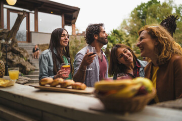 Group of friends at picnic sharing laughter and drinks at a rustic backyard table, enjoying a...