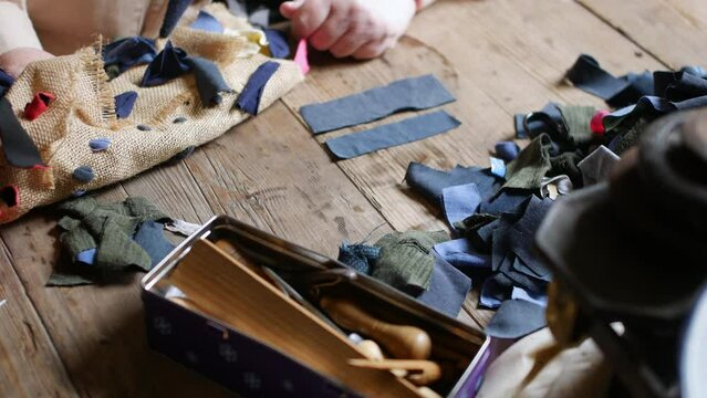 Close-up of hand working with textile pieces on a wooden table, alongside sewing tools. Detailed view of fabric rag rug crafting, ideal for handwork and DIY concepts