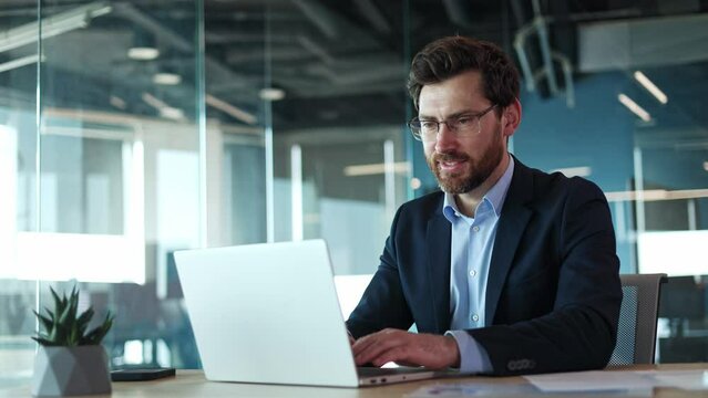 Smiling caucasian male wearing glasses with protective coating and typing on portable computer on background of office environment. Cheerful young businessman doing favorite work with pleasure.