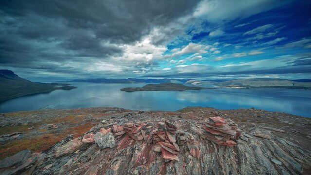 Whirling stormy clouds are backlit by the setting sun above the serene fjord and withered mountains in the time-lapse video.