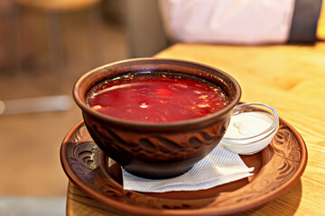 Hearty portion of classic borscht in carved clay bowl with sour cream in glass bowl, conveys homely atmosphere of Eastern European cuisine on wooden background