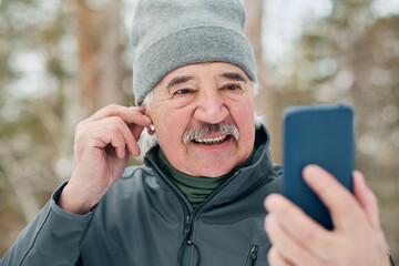 Medium closeup of cheerful senior man fixing wireless earphone while talking on video call