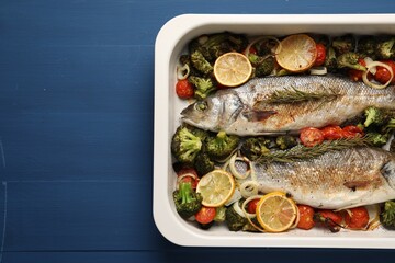 Delicious fish with vegetables and lemon in baking dish on blue wooden table, top view. Space for text