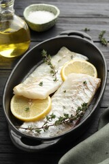 Fresh raw cod fillets with thyme and lemon in baking dish on black wooden table