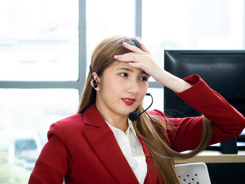Asian female customer service representative in a red blazer looking exhausted and stressed while at her office workstation
