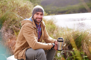 Camping, portrait and man with coffee by lake in nature for adventure, travel and outdoor vacation. Holiday, smile and male person with hot beverage for relax, peace and winter morning in countryside