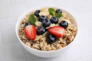 Tasty oatmeal with strawberries, blueberries and almond petals in bowl on white tiled table, closeup