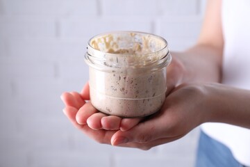 Woman holding glass jar with fresh sourdough starter on blurred background, closeup. Space for text