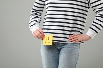 Cystitis. Woman holding sticky note with drawn sad face on grey background, closeup. Space for text