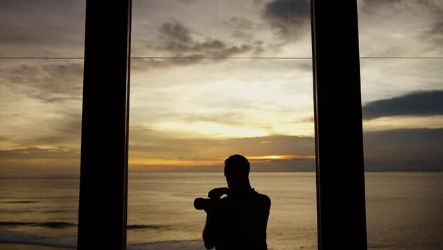 Silhouette of a man taking photos of sunset at the sea through window. Unrecognizable talented photographer with camera shooting the ocean view. Hobby and lifestyle
