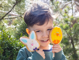 cute little baby boy with cookies on stick.toddler child is smiling.easter cookies bunny, rabbit shape drawing.spring time,outdoor environment, green bush.