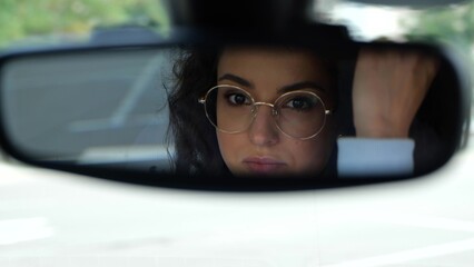 close up of young woman looking at rearview mirror and touching her hair while driving a car....