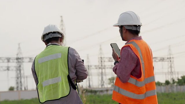 Two men wearing safety vests and hard hats are standing in a field. One of them is holding a walkie talkie